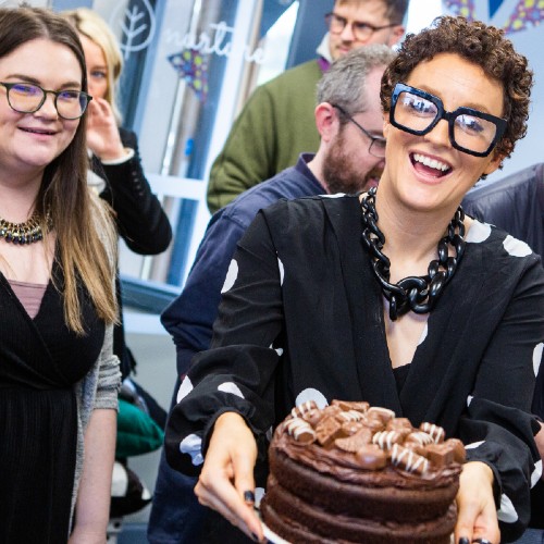 A group of people at a Big Cuppa fundraising event, with the host carrying a large chocolate cake with chocolates on top.
