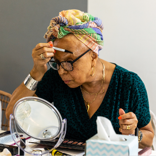 A women in a colourful headscarf applying make up in a mirror.