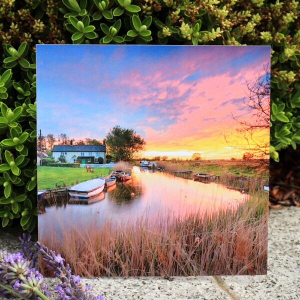 A greetings card of a broadland scene at sunrise, with boats, reeds, a house and calm waters